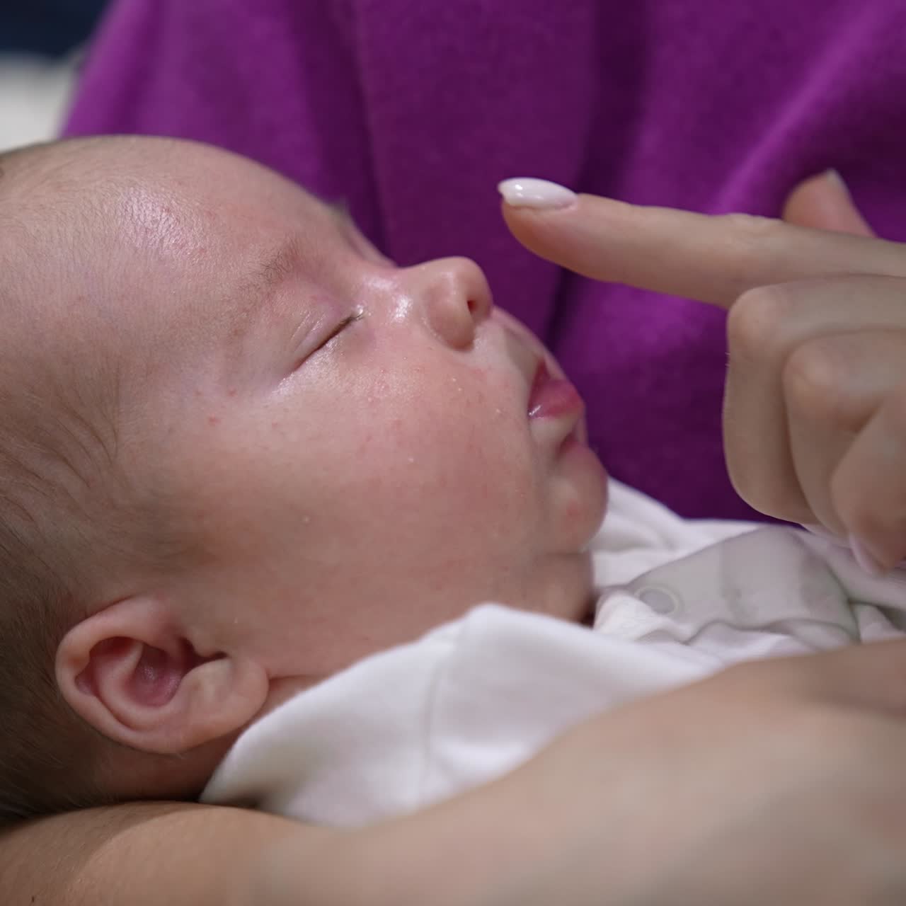 Beautiful infant in mother's embraces sleeps sweetly. Mother is trying to wake her son up by touching his chin, nose, cheeks. Cute boy doesn't want to wake up