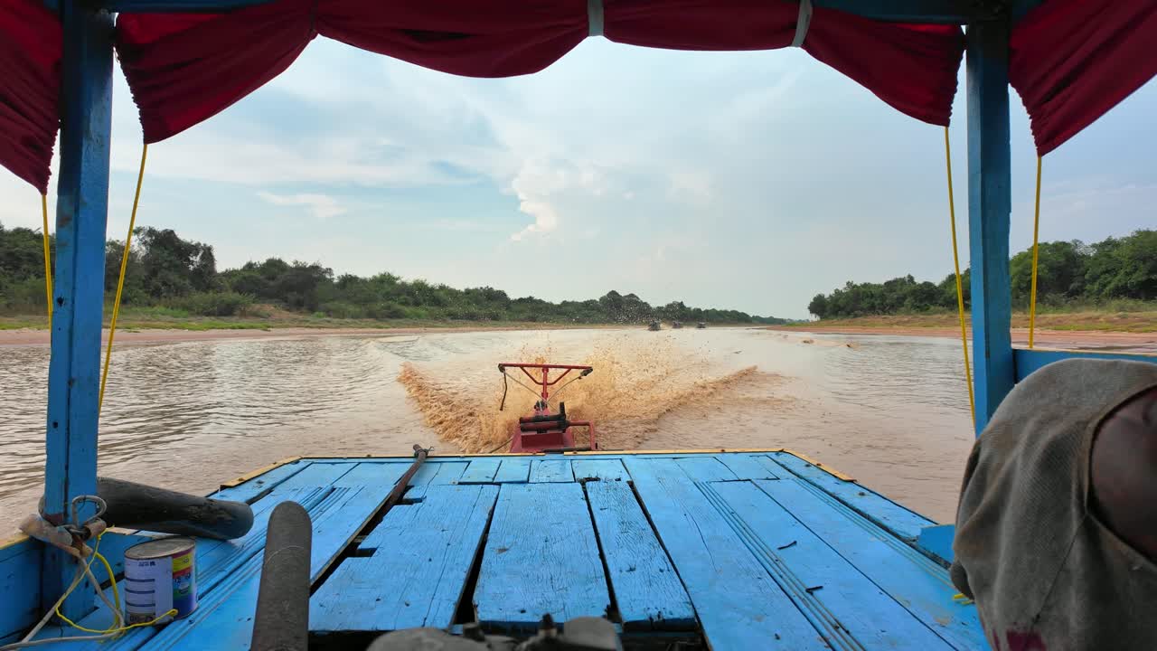 A traditional, colourful Cambodian boat moves through the muddy waters of Bassac River. Its long-tail engine churns, cutting through the current as the riverbank stretches in the background.