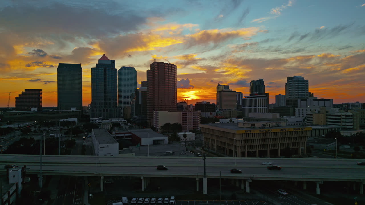 Aerial descend of downtown Tampa skyline glowing at sunset with colorful clouds and cars driving on highway