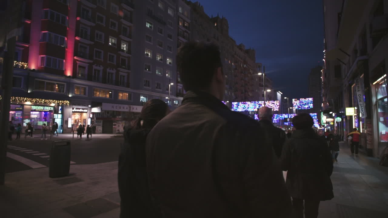 Panning slow motion shot following various people down the street in Madrid. Illuminated area at night.