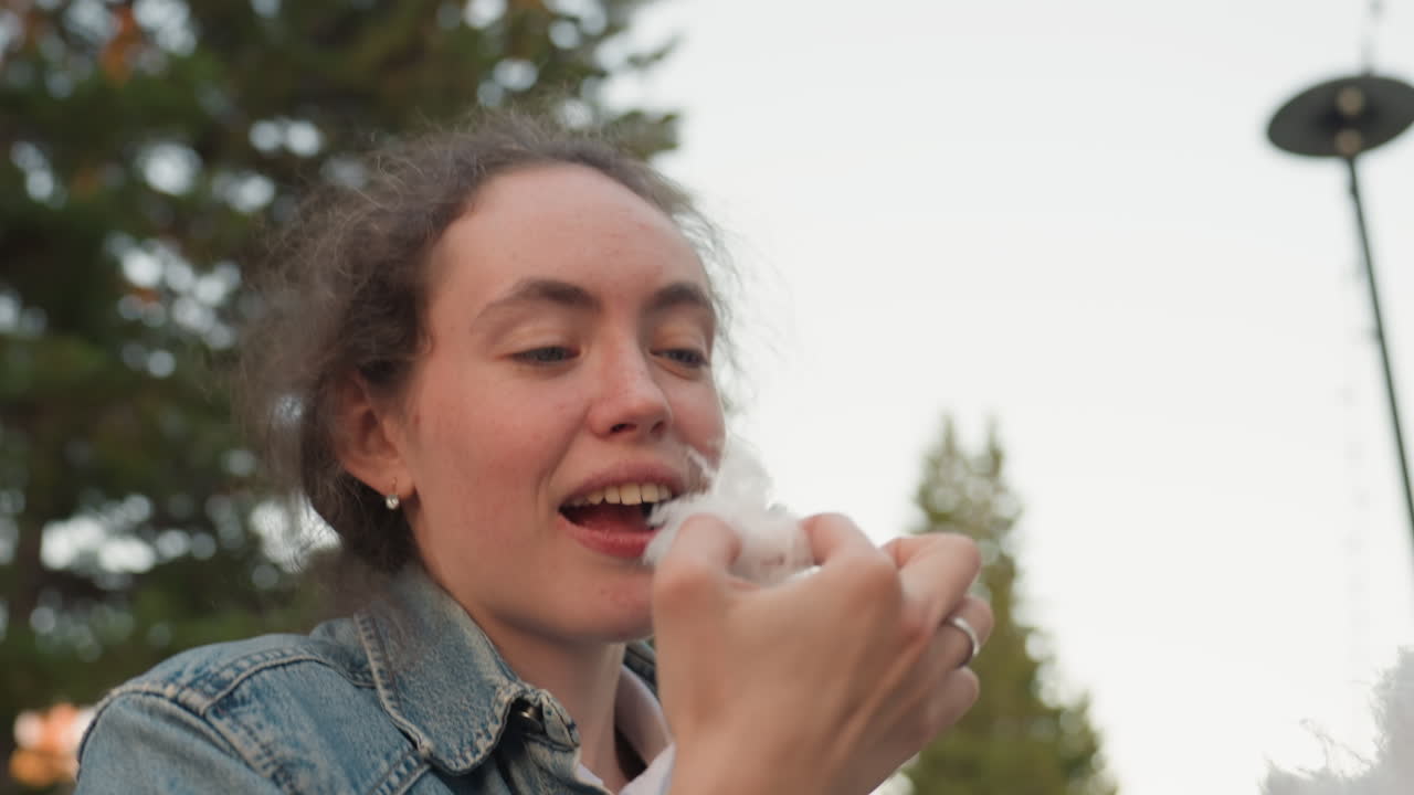 White woman tasting cotton candy outdoors, closeup denim jacket, pine trees background, evening light, gentle pull of fluffy piece, savoring texture and sugar, thoughtful expression, candid festival