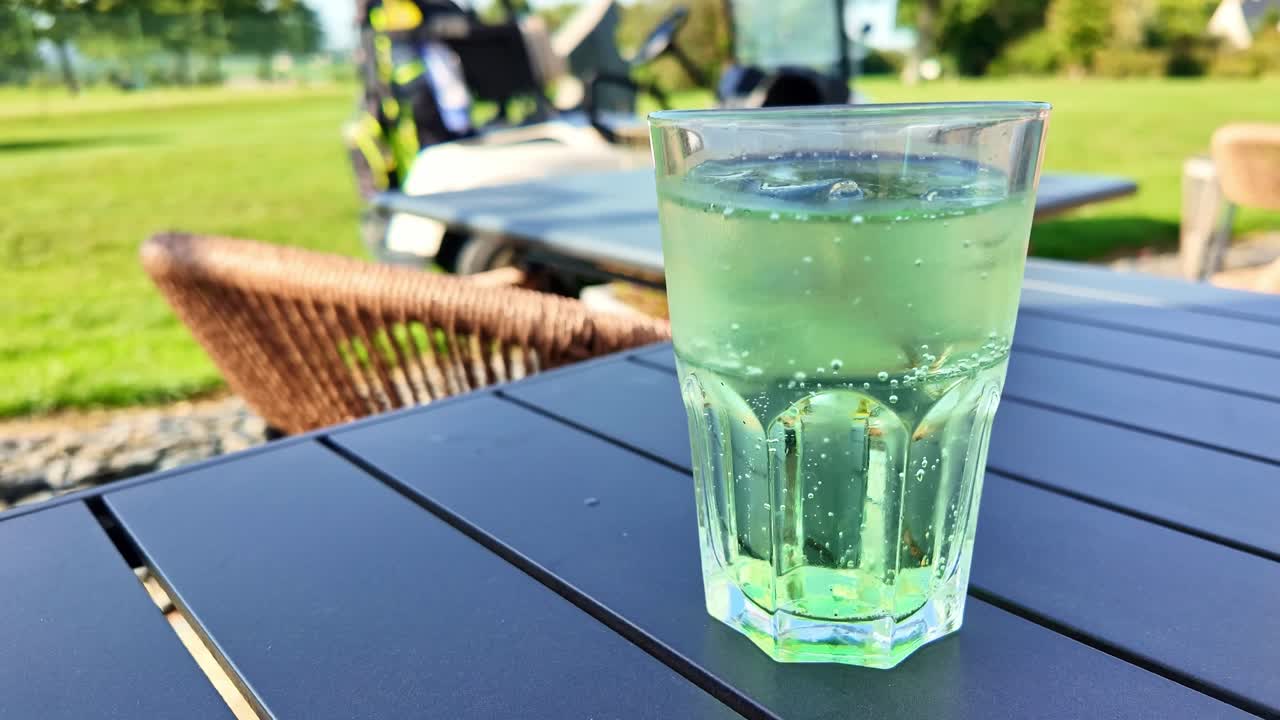 Glass of refreshing green diabolo drink on outdoor table, golf course and golf cart in background on sunny day. Close-up