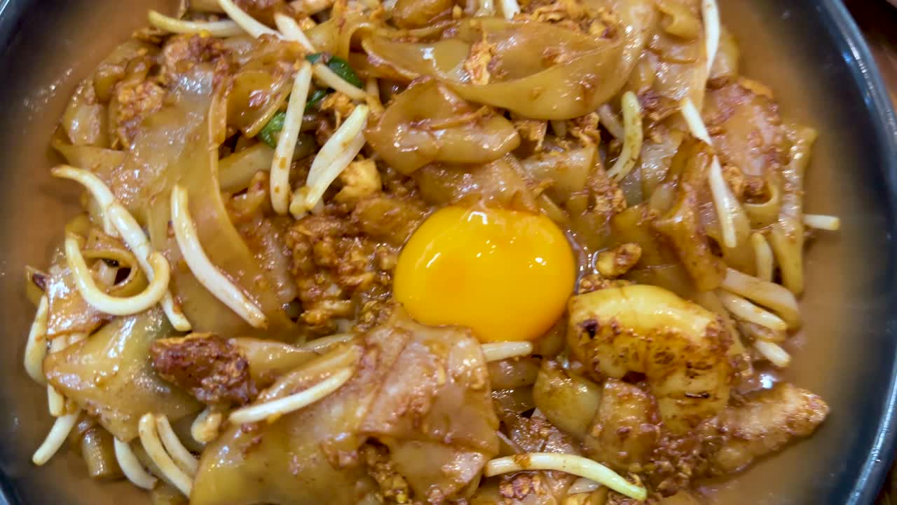 Overhead shots of assorted Singaporean dishes including fried noodles, turnip cake, dim sum, and rice rolls, under bright indoor lighting with smooth camera panning