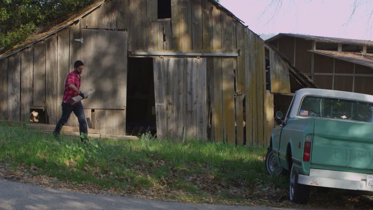 A man carries a board to a pickup truck parked outside an old barn