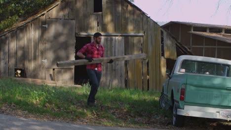 a man carries a board to a pickup truck parked outside an old barn