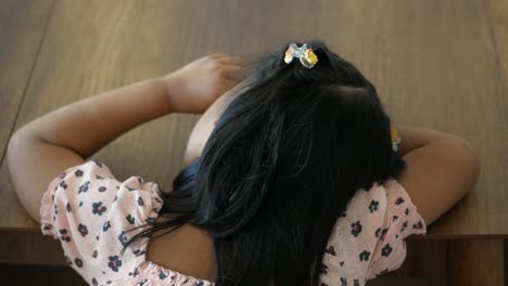 a young girl with long dark hair wearing a pink floral dress sits at a wooden table, looking away from the camera.