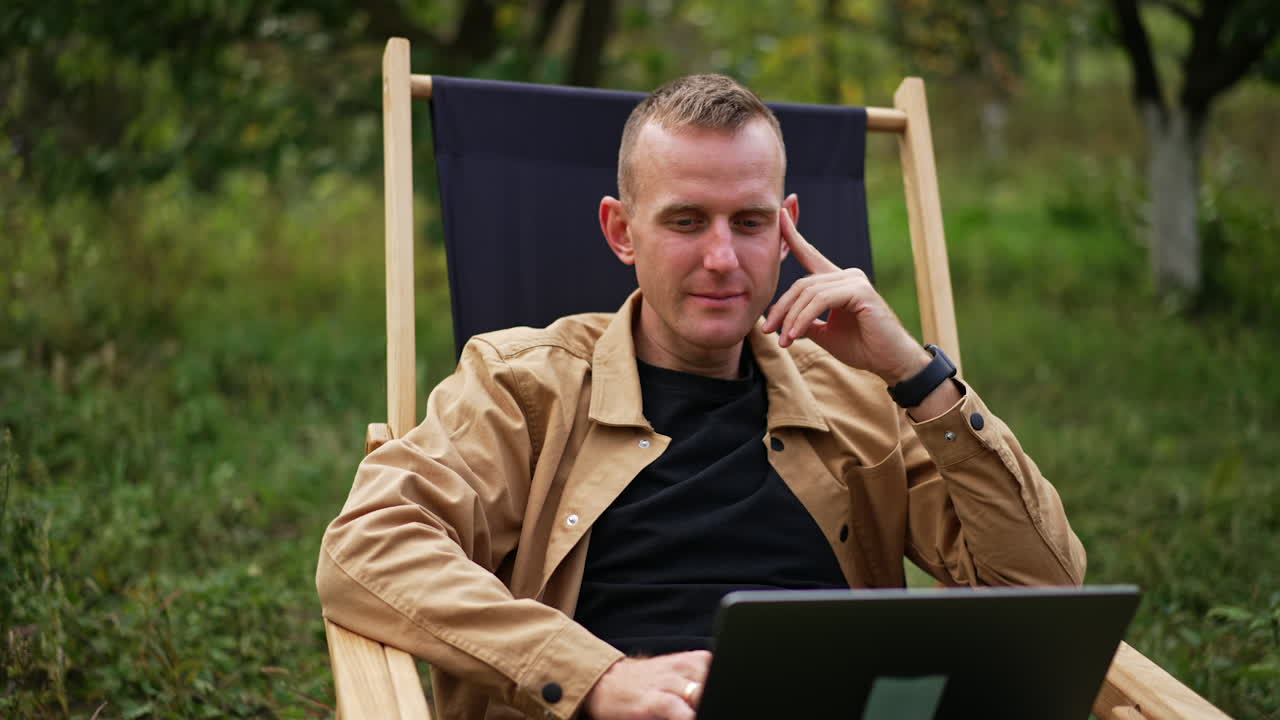 Pensive sitting in garden chair looks thoughtfully at his laptop screen. Freelancer working in nature has found some mistake and looks closer at computer.