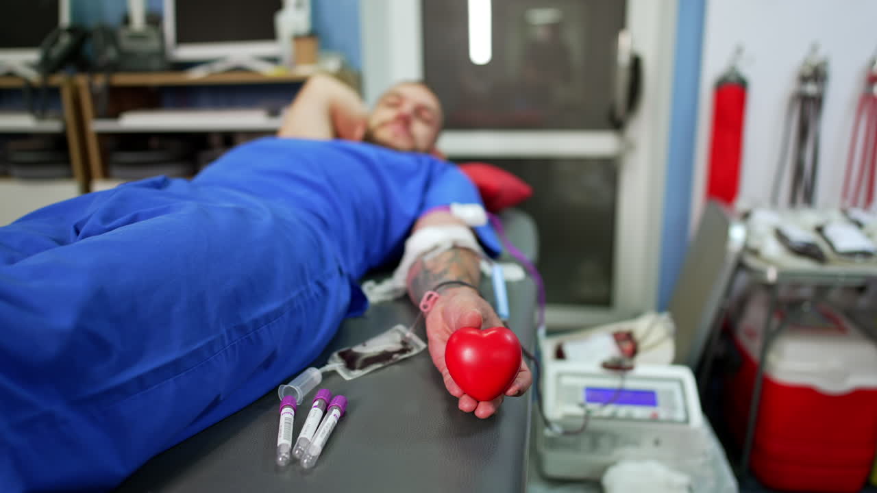 Unrecognized man wearing blue clothes lies on the couch holding a red heart in hand. Male is giving his blood in the hospital. Blood donation concept.