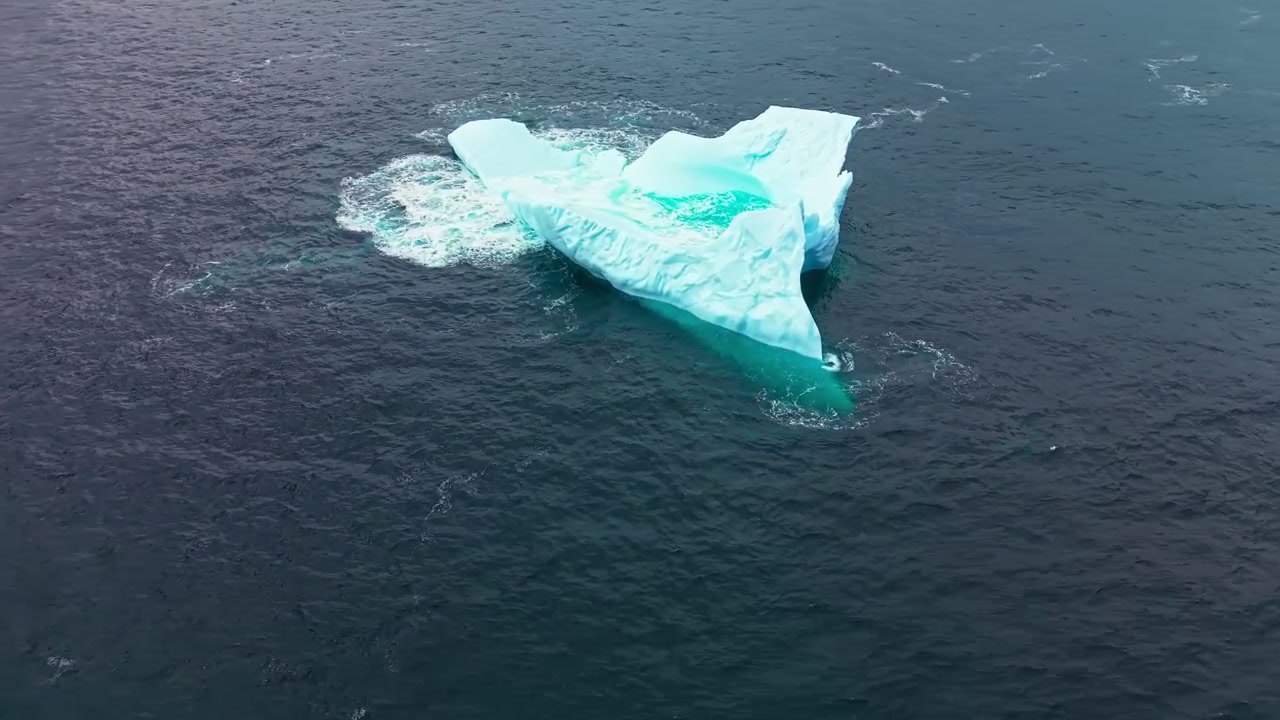 Drohnenaufnahme von oben eines kompakten Eisbergs, umgeben von sonnenbeschienenem, offenem Atlantikwasser.