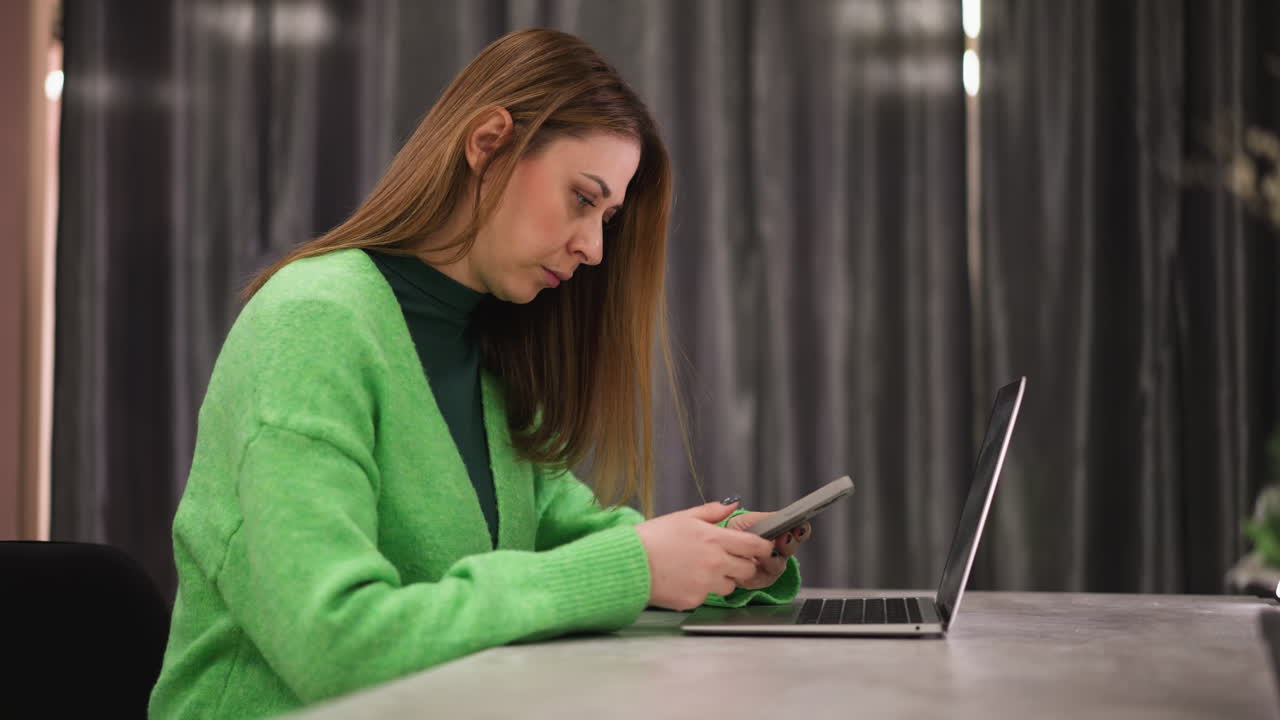 First-person perspective of a woman wearing a green sweater, sitting at a table with a laptop in front of her, while she uses her phone