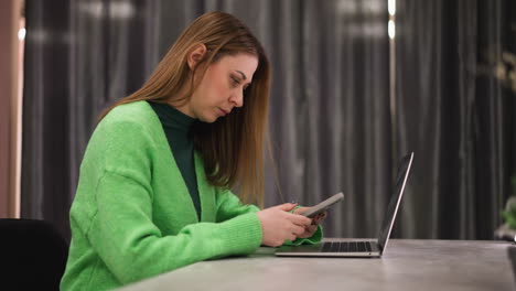 perspectiva en primera persona de una mujer con un suéter verde, sentada en una mesa con una computadora portátil delante de ella, mientras usa su teléfono