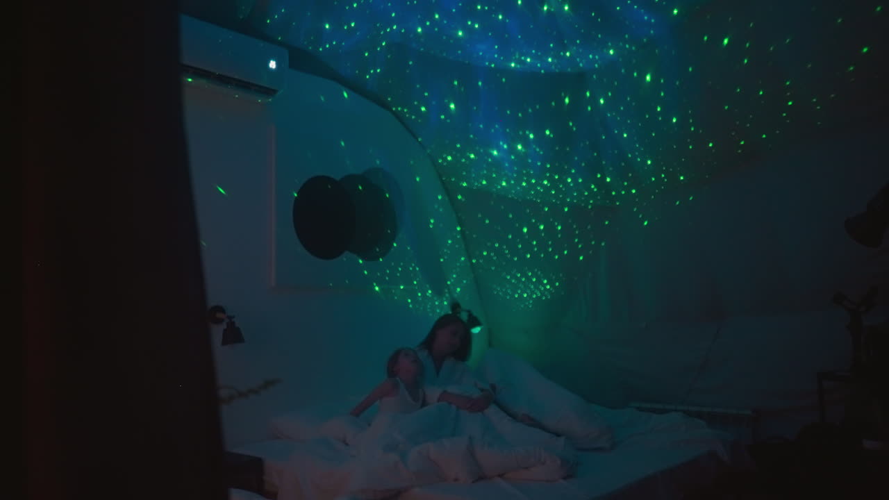 mujer con hijos mira el cielo estrellado brillante que brilla en el techo sentada en la cama en una habitación moderna. fuente de luz tenue haciendo la iluminación en la premisa suave y relajante cámara lenta