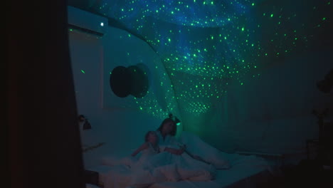 woman with children looks at bright starry sky glowing on ceiling sitting on bed in modern room. dim light source making lighting in premise soft and soothing slow motion