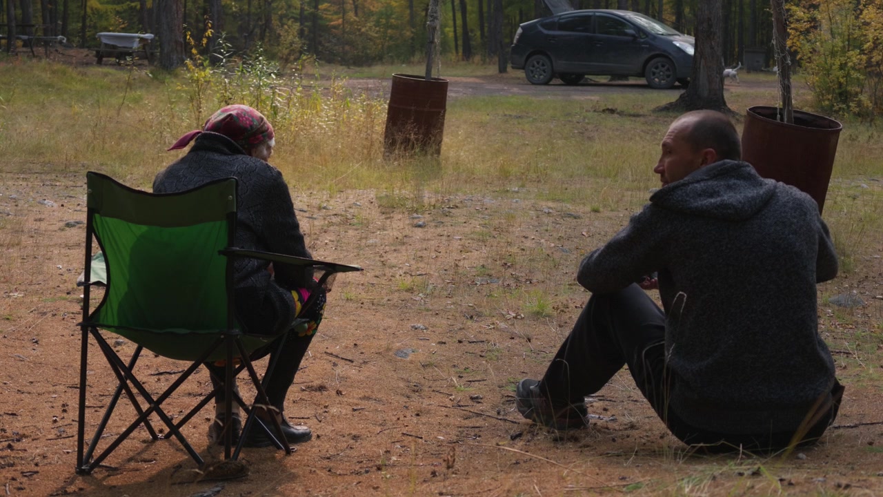 Two individuals converse while seated in a wooded area, one in a green folding chair and the other on the ground, with a parked car and rusted metal barrels visible behind them.