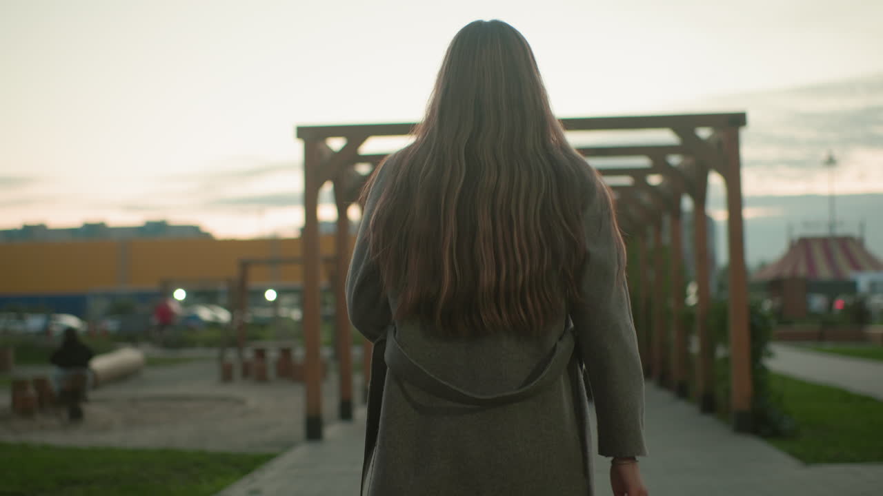 Back view of woman sitting on wooden bench outdoors holding book in hand, wearing grey coat, long hair flowing down, peaceful evening atmosphere with pergola structure and soft sunset in background