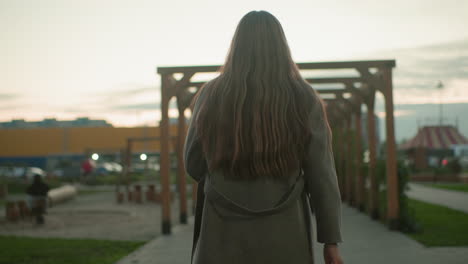 back view of woman sitting on wooden bench outdoors holding book in hand, wearing grey coat, long hair flowing down, peaceful evening atmosphere with pergola structure and soft sunset in background