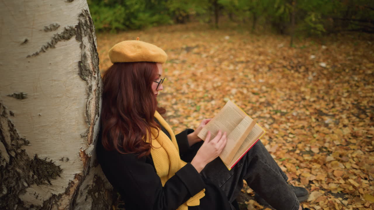 estudiante con boina amarilla y bufanda se sienta al aire libre apoyándose en un árbol, repasando pensativamente las páginas de los libros, rodeada de hojas doradas de otoño, parece involucrada en una lectura pacífica y reflexiva