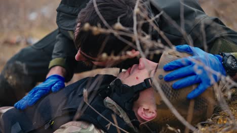 Nahaufnahme:-Ein-Brünetter-Mann-Mit-Bart-In-Dunkelgrüner-Uniform-Und-Blauen-Medizinischen-Handschuhen-Lauscht-Der-Atmung-Eines-Verwundeten-Soldaten-In-Tarnuniform-Und-Körperpanzerung,-Der-Während-Kampfhandlungen-Auf-Dem-Boden-In-Der-Steppe-Liegt