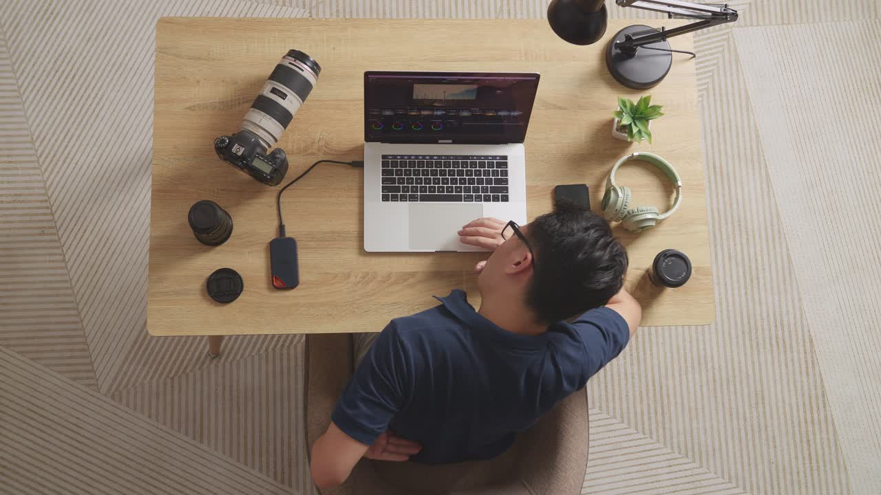 Top View Of Asian Male Color Grading Having A Backache While Sitting In The Workspace Using A Laptop Next To The Camera Editing The Video At Home