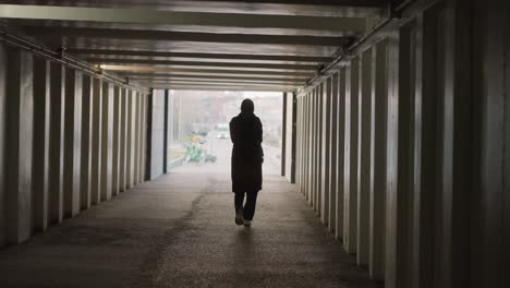 a back shot of a girl wearing a backpack, slowly walking through an underpass or tunnel. the tunnel has a series of vertical support beams, and somber mood as she approaches the exit