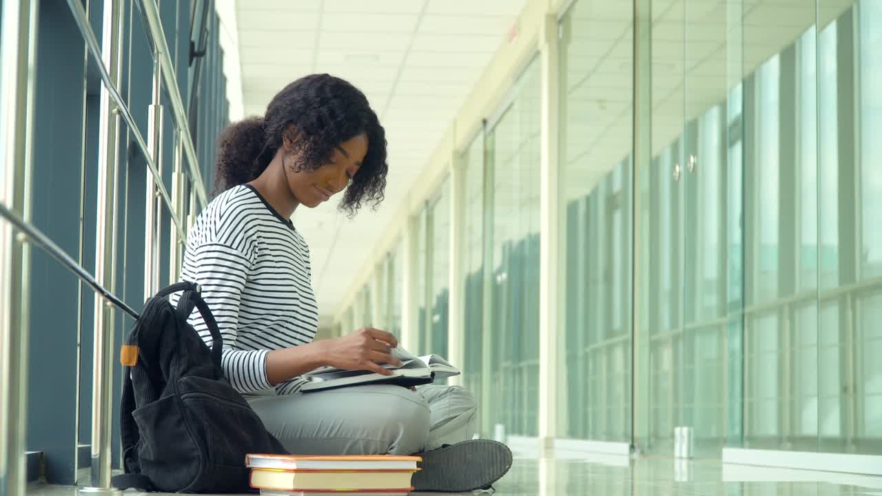 African american woman student sitting on the floor with a books in the university coridor. New modern fully functional education facility. Concept of online education