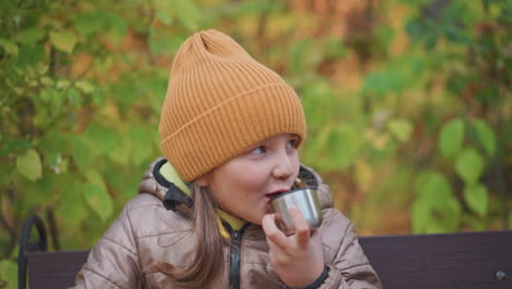 young girl wearing brown puffer jacket and orange knit beanie drinks from small metal cup and licks lips, sitting relaxed on bench against colorful autumn foliage in peaceful forest setting