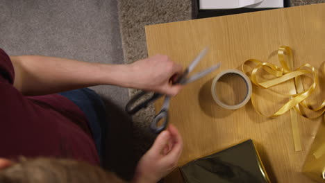 overhead shot of man using sticky tape and ribbon to gift wrapped present on table at home