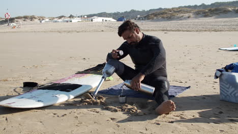 toma estática de un surfista masculino con pierna artificial sentado en la orilla arenosa y sirviendo té del termo