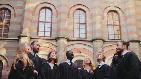 Grupo-De-Graduados-Multiétnicos-Vestidos-Con-Ropa-Tradicional-Lanzando-Sus-Gorras-Al-Aire-Alegremente-Frente-A-La-Universidad