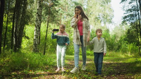 mother walks with children through green forest trail, holding boy hand while pointing ahead, girl walks beside filming surroundings with camera, boy appears upset during peaceful woodland stroll