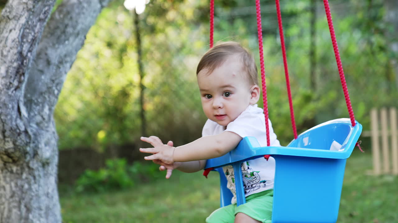 Caucasian baby boy swaying in the blue swing. Lovely child enjoying summertime in the nature. Blurred background.