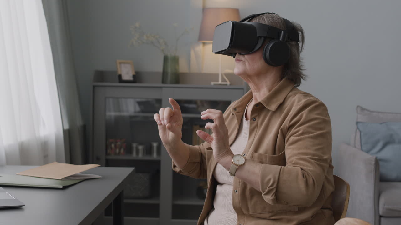 пожилая женщина использует очки гарнитуры виртуальной реальности и двигает руками, сидя на стуле в современной гостиной 1