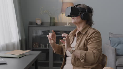 mujer mayor que usa gafas de realidad virtual y mueve las manos mientras se sienta en una silla en una sala de estar moderna 1