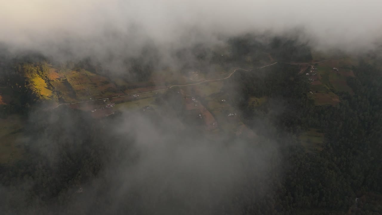 High drone shot moving forward through dark clouds and revealing the rural Zacatlán valley below, with fields, scattered houses, forest areas and a winding road