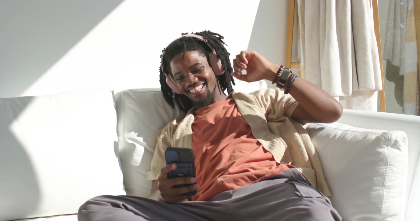 Uomo afroamericano sdraiato su un divano bianco con cuffie rosa, che reagisce a un avviso del telefono, sorridendo