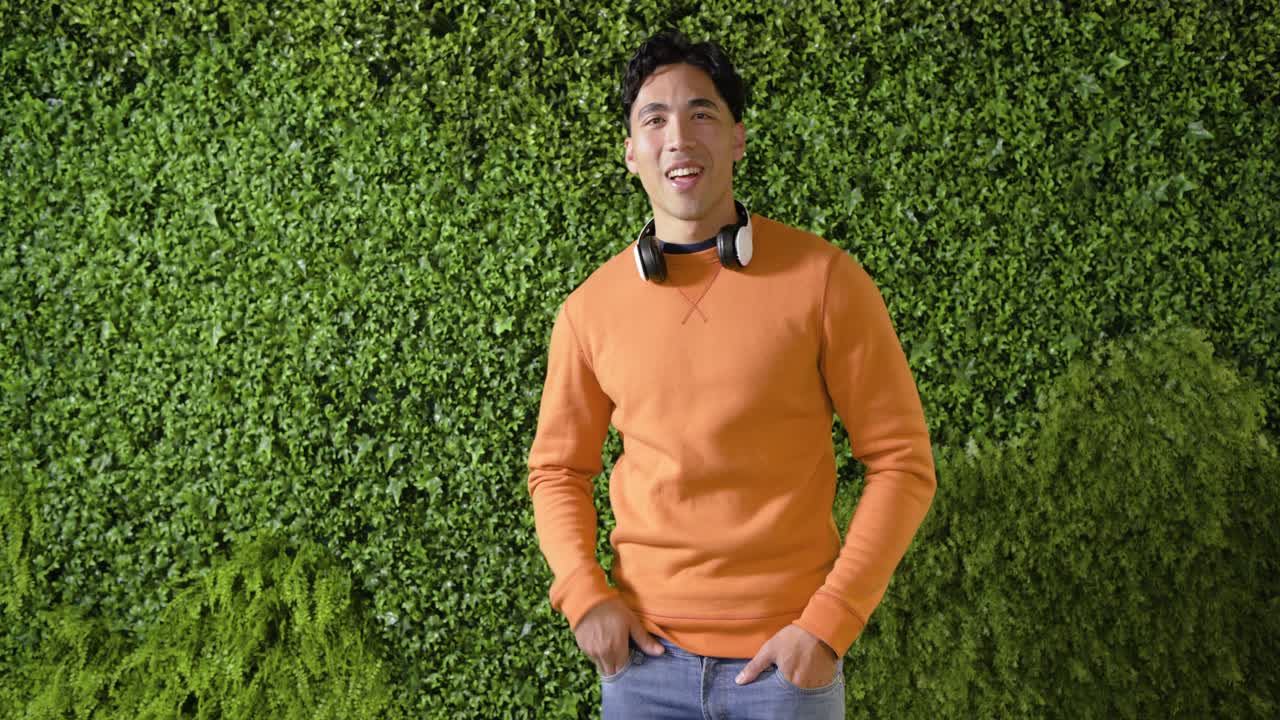 生い茂る緑の壁の前で、カメラに向かって顎を上げながら微笑み、身振り手振りを交えて話す男性。