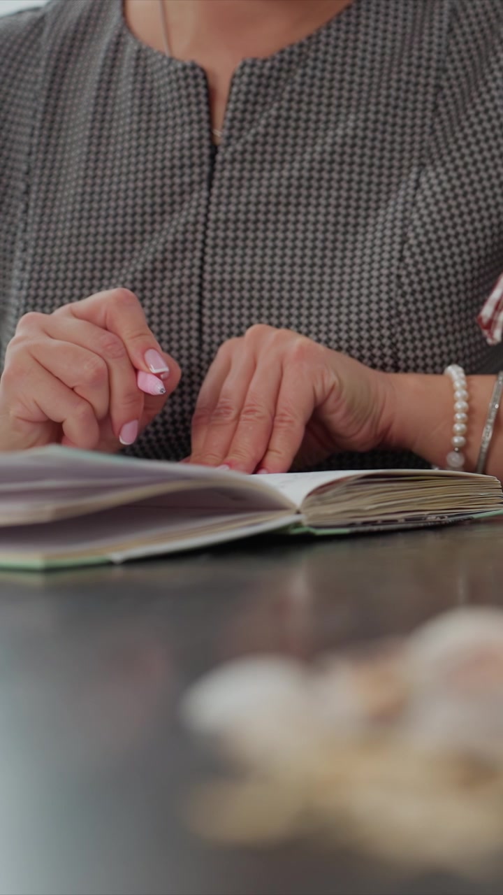 Primo piano di una donna che scrive su un quaderno aperto con una penna rosa, indossando abiti fantasia e gioielli. La scena mostra le mani con il quaderno su una superficie piana, catturando un'attività concentrata.