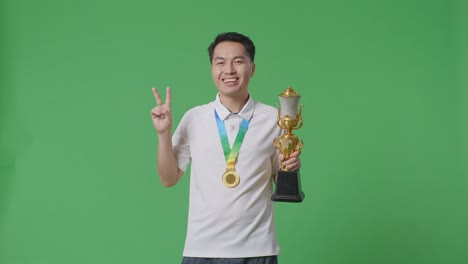 asian man with a gold medal and trophy showing peace gesture and smiling to camera as the first winner on green screen background in the studio