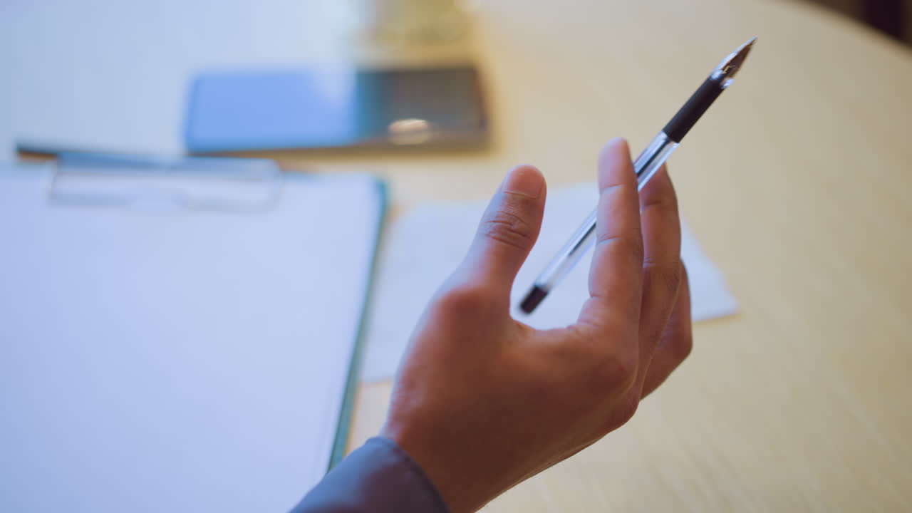 Close-up of man holding pen over clipboard and paper on table in bright indoor setting, suggesting readiness to write or take notes, with smartphone and glass visible in softly blurred background
