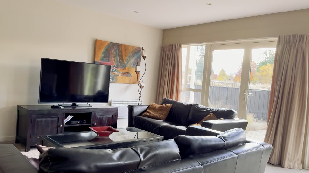 Camera smoothly moves from kitchen island into a bright, modern living room with large windows, leather sofas, TV, and contemporary decor. Natural daylight fills the space