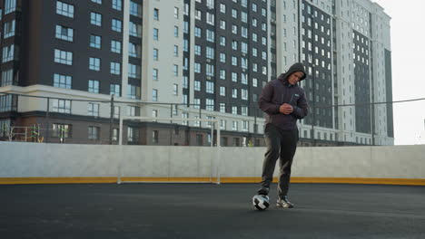 hombre de pie en el campo de deportes con un pie en la pelota de fútbol, manos juntas, y girando la cabeza, rodeado de edificios modernos de la ciudad bajo la luz del día brillante