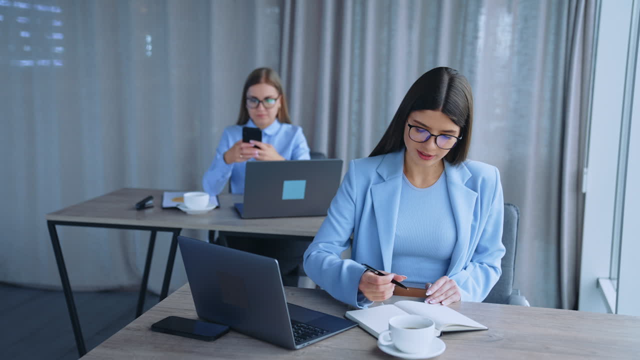Female employees sitting at desks in the office. Brunette colleague writing in the notebook and blonde lady looking at her phone.