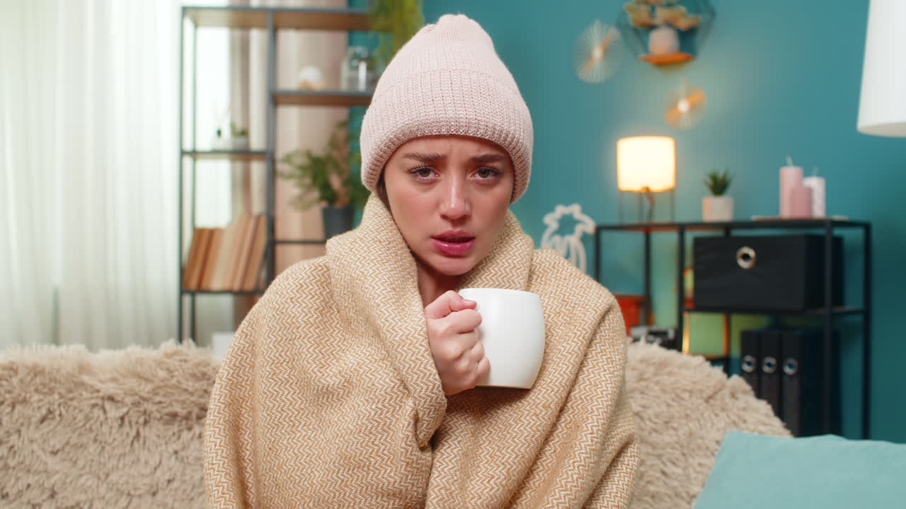 Young woman drinking hot tea in cozy blanket wearing hat feeling cold and sick resting on sofa today