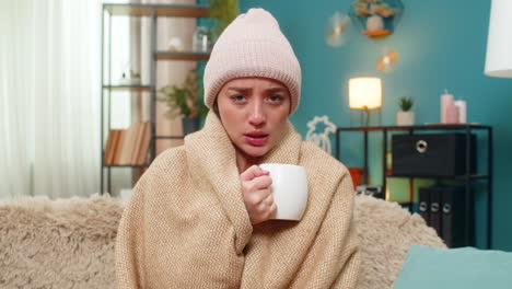 young woman drinking hot tea in cozy blanket wearing hat feeling cold and sick resting on sofa today