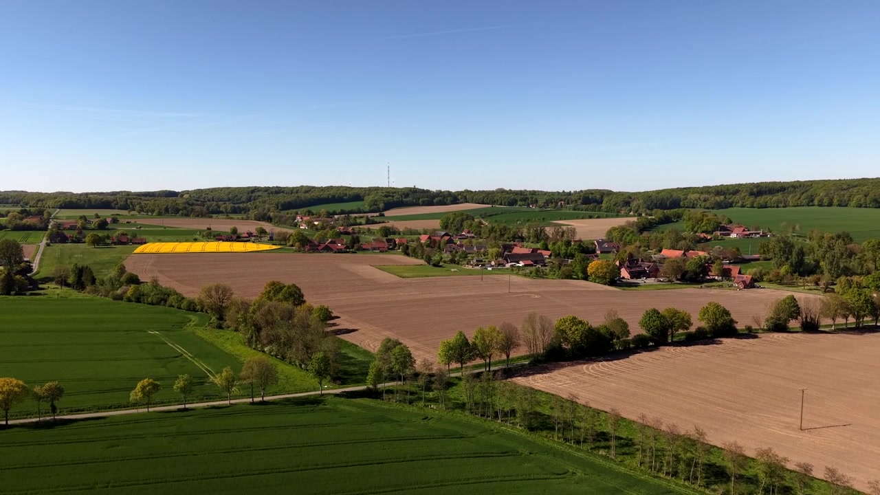Glooiend landbouwgebied en een lappendeken van velden met een klein Europees dorp en met bomen omzoomde wegen onder een helderblauwe hemel, met idyllische lentekleuren en een landelijk plattelandslandschap. Breed opstijgend luchtbeeld.