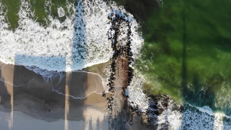 descending birds eye view of a rock jetti that sits between a lime green, darkened blue ocean that crashes waves into dark, slippery rocks amongst the pale, coarse sand shaded by afternoon shadows