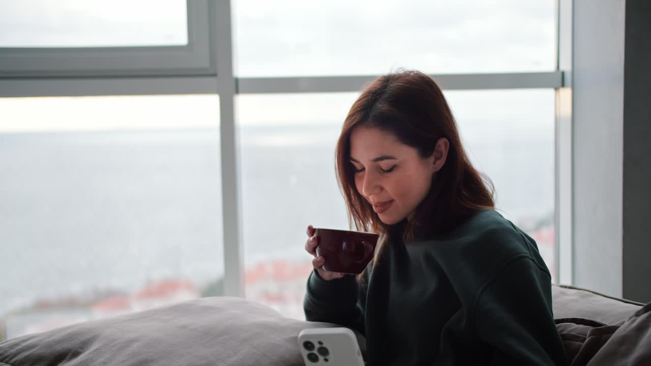 어두운 녹색 재을 입은 행복한 갈색 여성이 갈색 컵에서 차를 마시며 바다를 내려다보는 현대 아파트에서 집에서 일하고 휴식을 취하는 동안 색 전화를 봅니다.