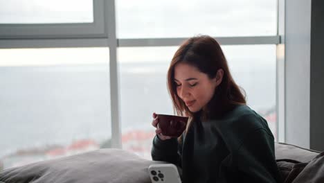 A-happy-brunette-woman-in-a-dark-green-jacket-drinks-tea-from-a-brown-cup-and-looks-at-a-white-phone-during-her-work-and-relaxation-at-home-in-a-modern-apartment-overlooking-the-sea