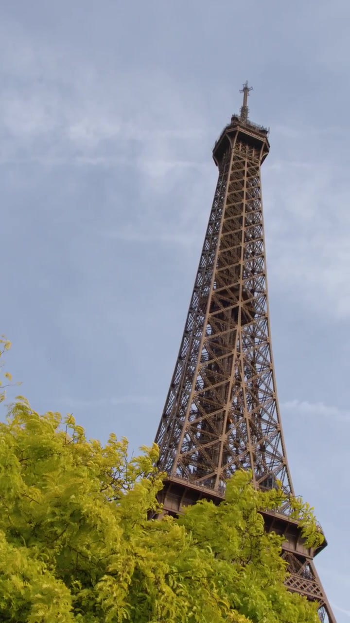 Una telecamera ad angolo basso esegue una panoramica a destra, rivelando l'iconica torre di ferro dietro un vivace fogliame verde.