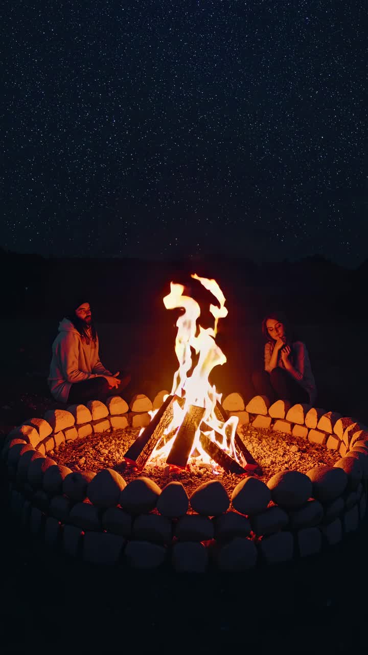 A low-angle video shot captures two people sitting by a campfire under a starry night sky